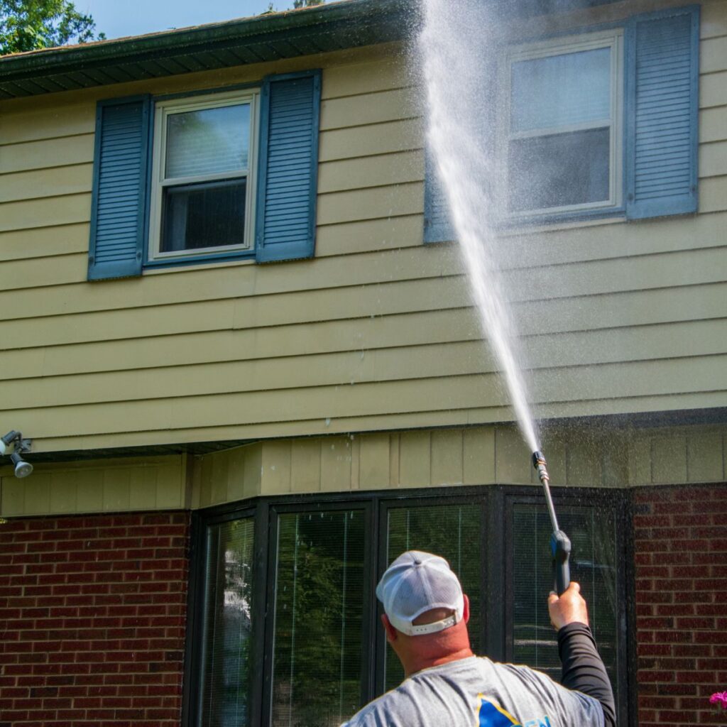 Worker soft washes the upper story of a house with blue shutters and beige siding