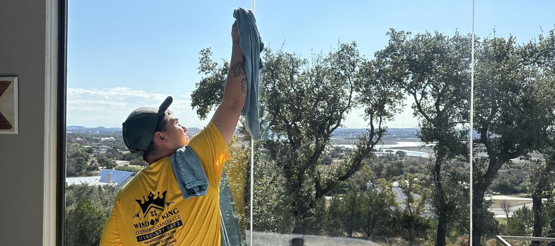 Window cleaning technician wipes down tall indoor windows with a towel, overlooking a scenic view