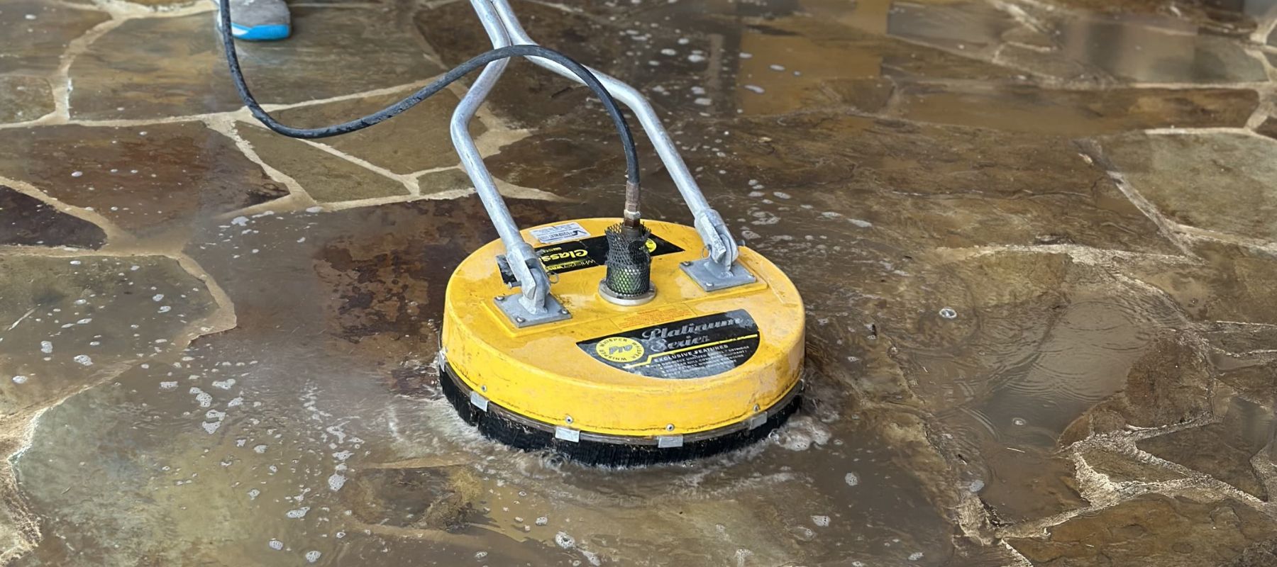 Yellow rotary surface cleaner scrubbing a stone patio covered in soapy water