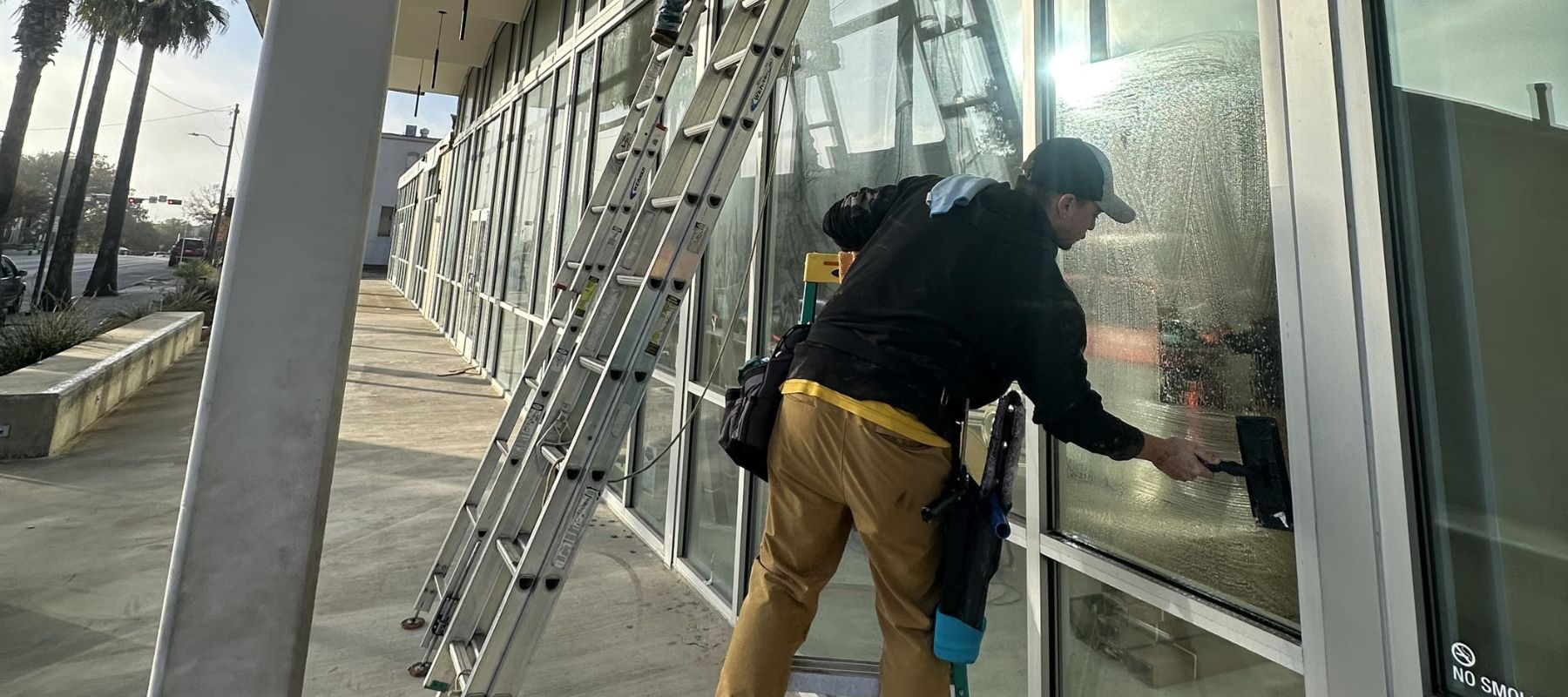 A man cleans tall exterior glass windows of a commercial building with ladders set up along the sidewalk