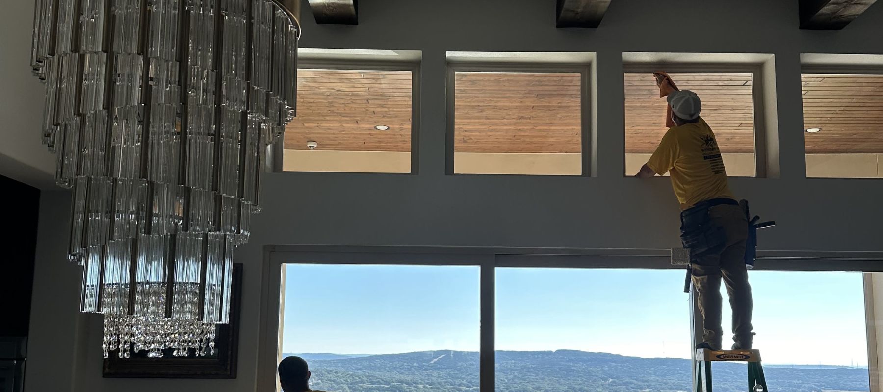 Technician on a ladder cleaning upper transom windows inside a home with a mountain view