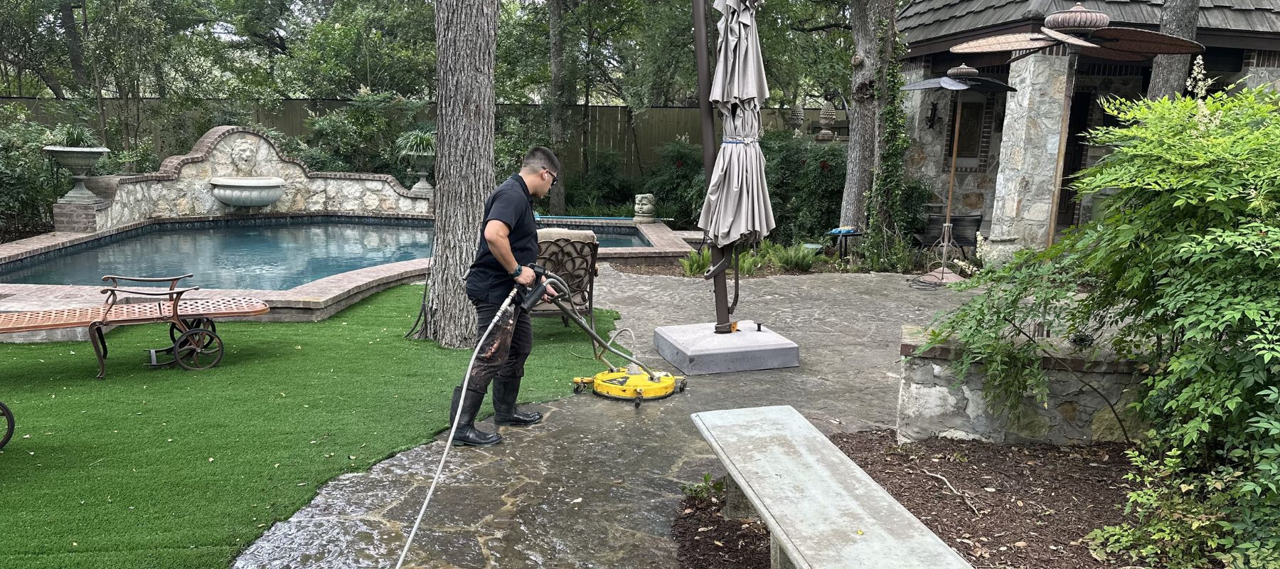 Worker power washing a stone patio beside a pool in a landscaped backyard.