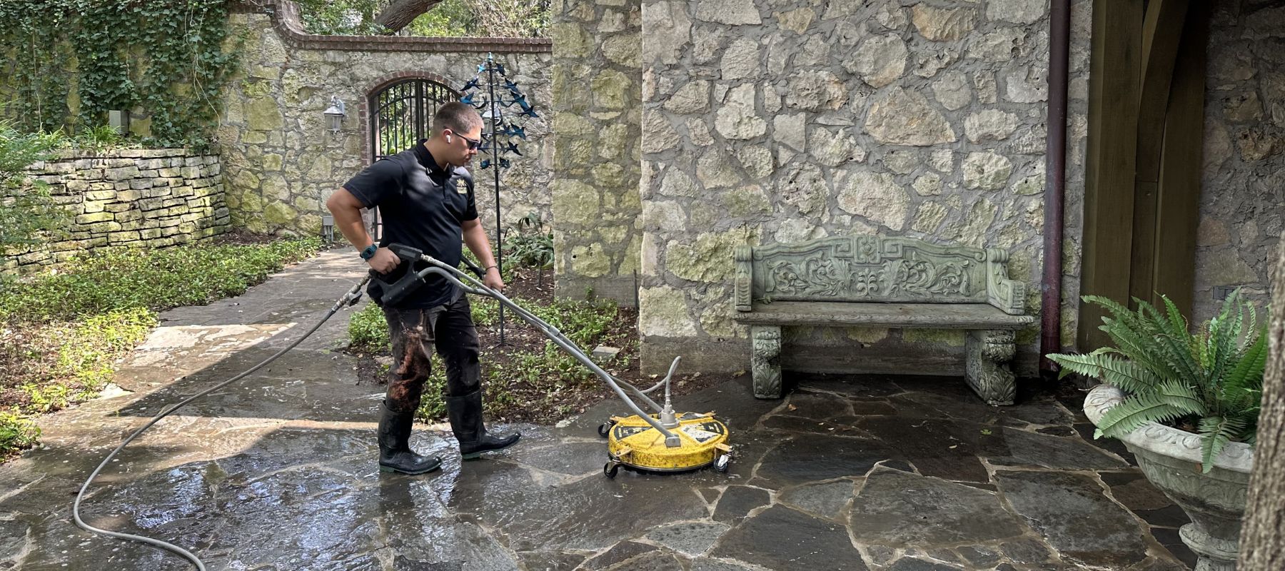 A worker operates a yellow surface cleaner to power wash a stone walkway near a stone wall and bench