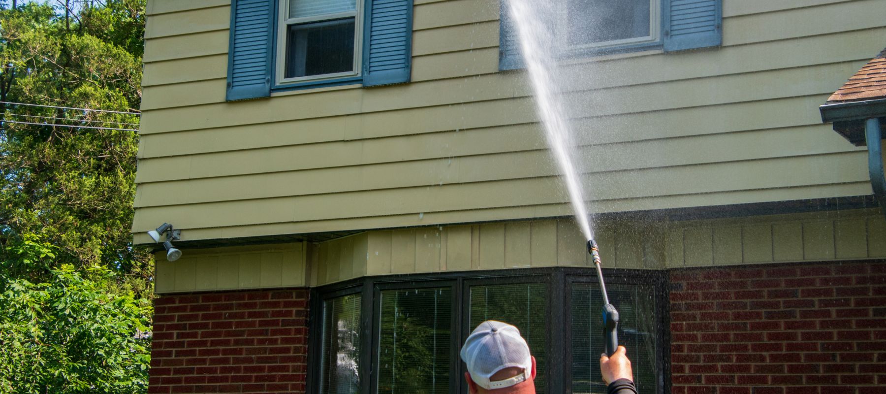 A man uses a high-pressure washer to clean the siding of a two-story house with blue shutters and red brick base