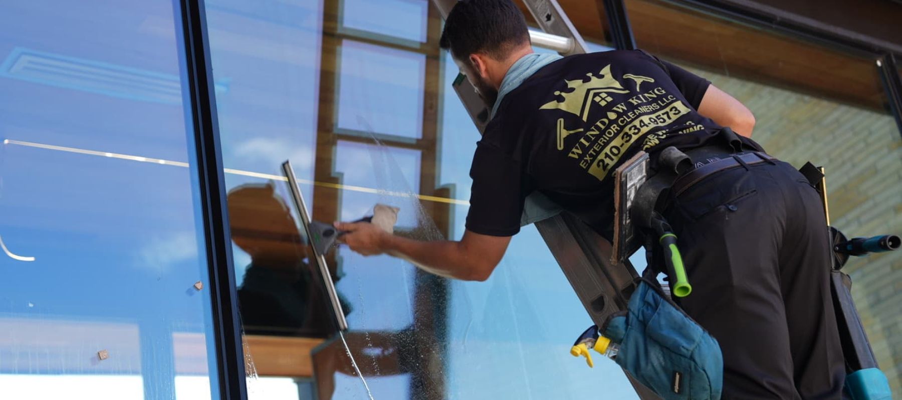 A professional window cleaner uses a squeegee to clean a large glass window while standing on a ladder