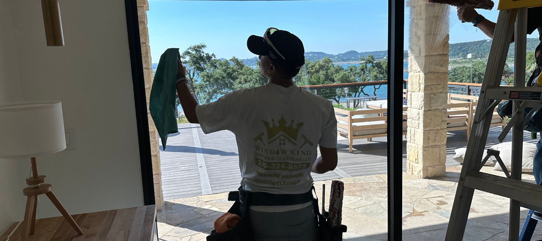 A professional window cleaner wipes a glass door overlooking a scenic lake and patio