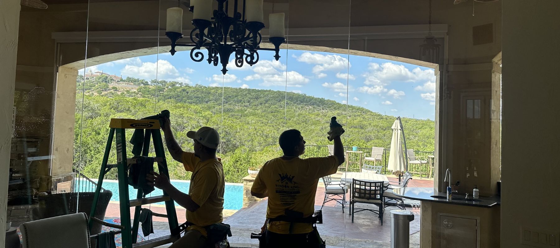Two window cleaners clean large glass doors inside a home with a view of a pool and hills
