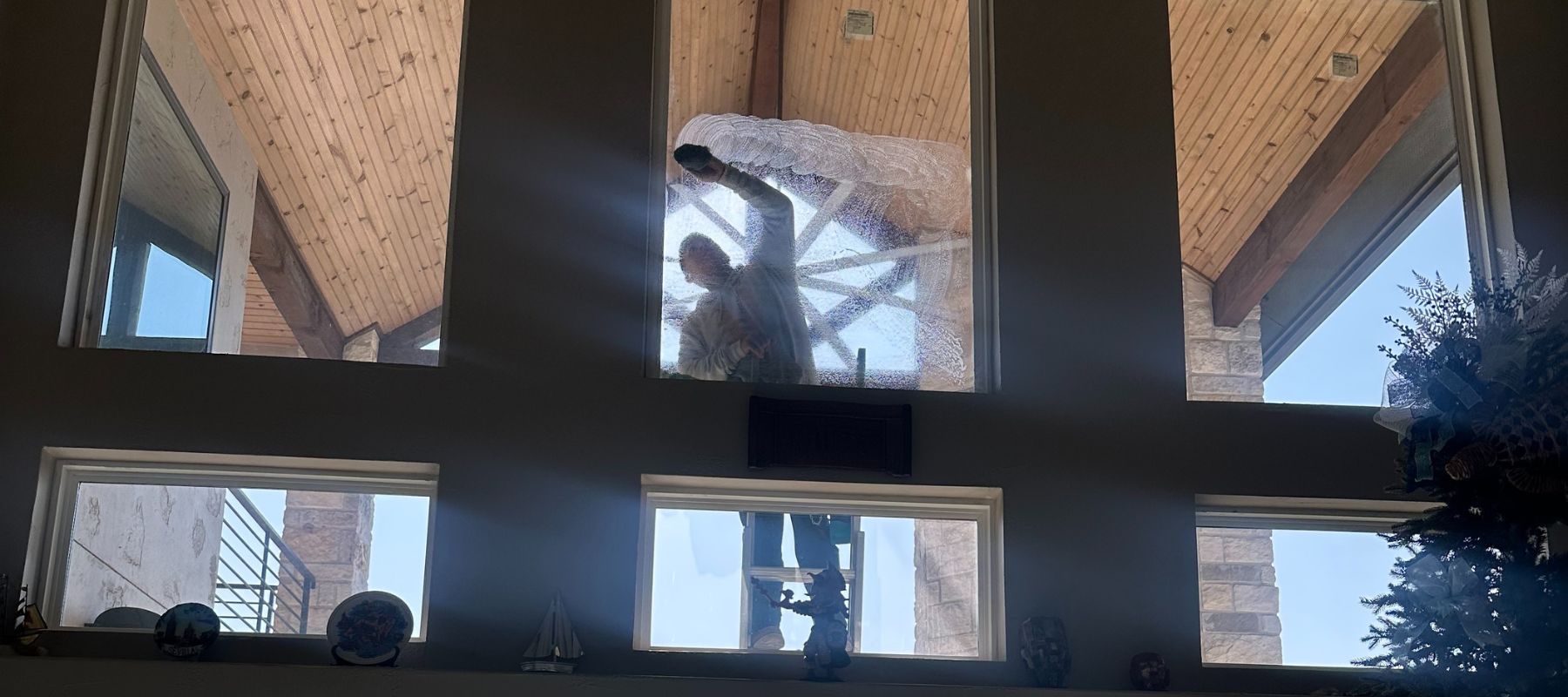 A person on a ladder is seen from inside as they scrub tall exterior windows covered in soap