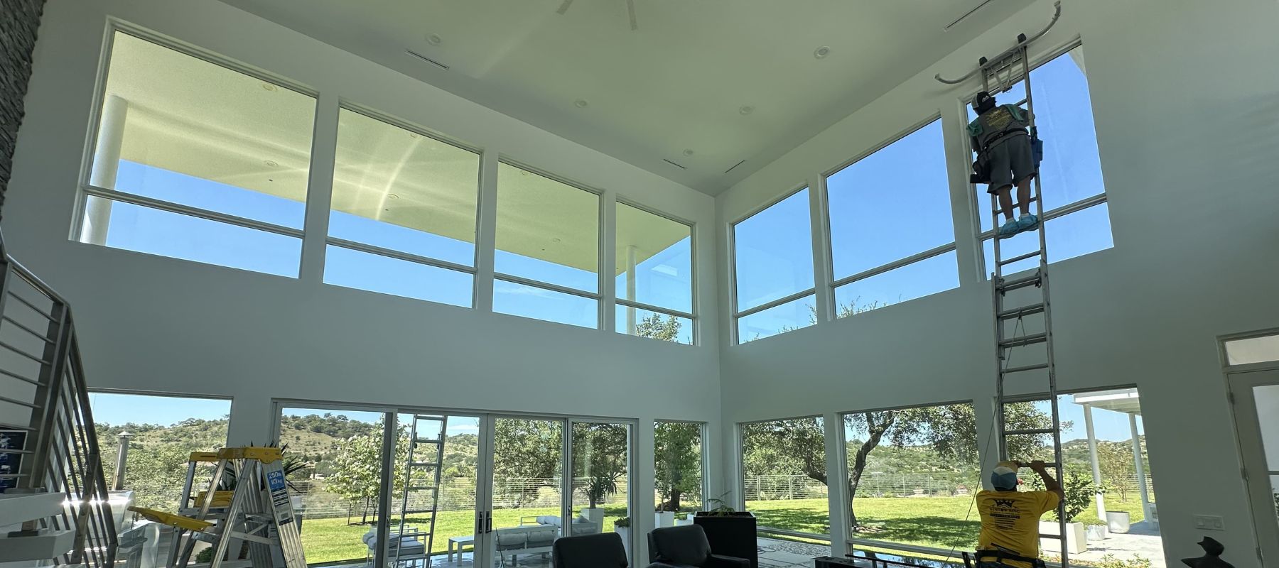 A window cleaner on a tall ladder cleans second-story windows in a bright living room with high ceilings